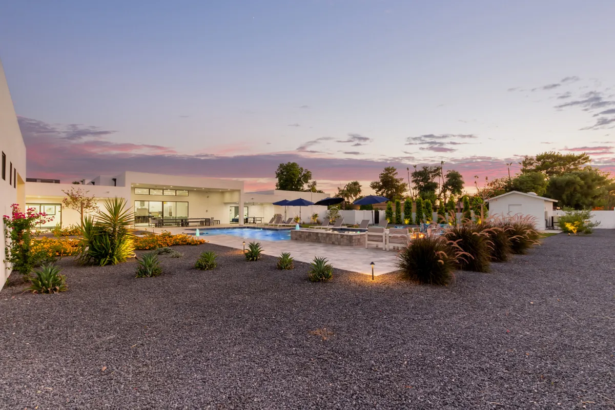 A tranquil backyard at sunset featuring a sparkling swimming pool, comfortable outdoor seating area, manicured gardens, and a vibrant sky. The scene captures a sense of privacy and luxury, ideal for hosting evening gatherings or relaxing after a night out.