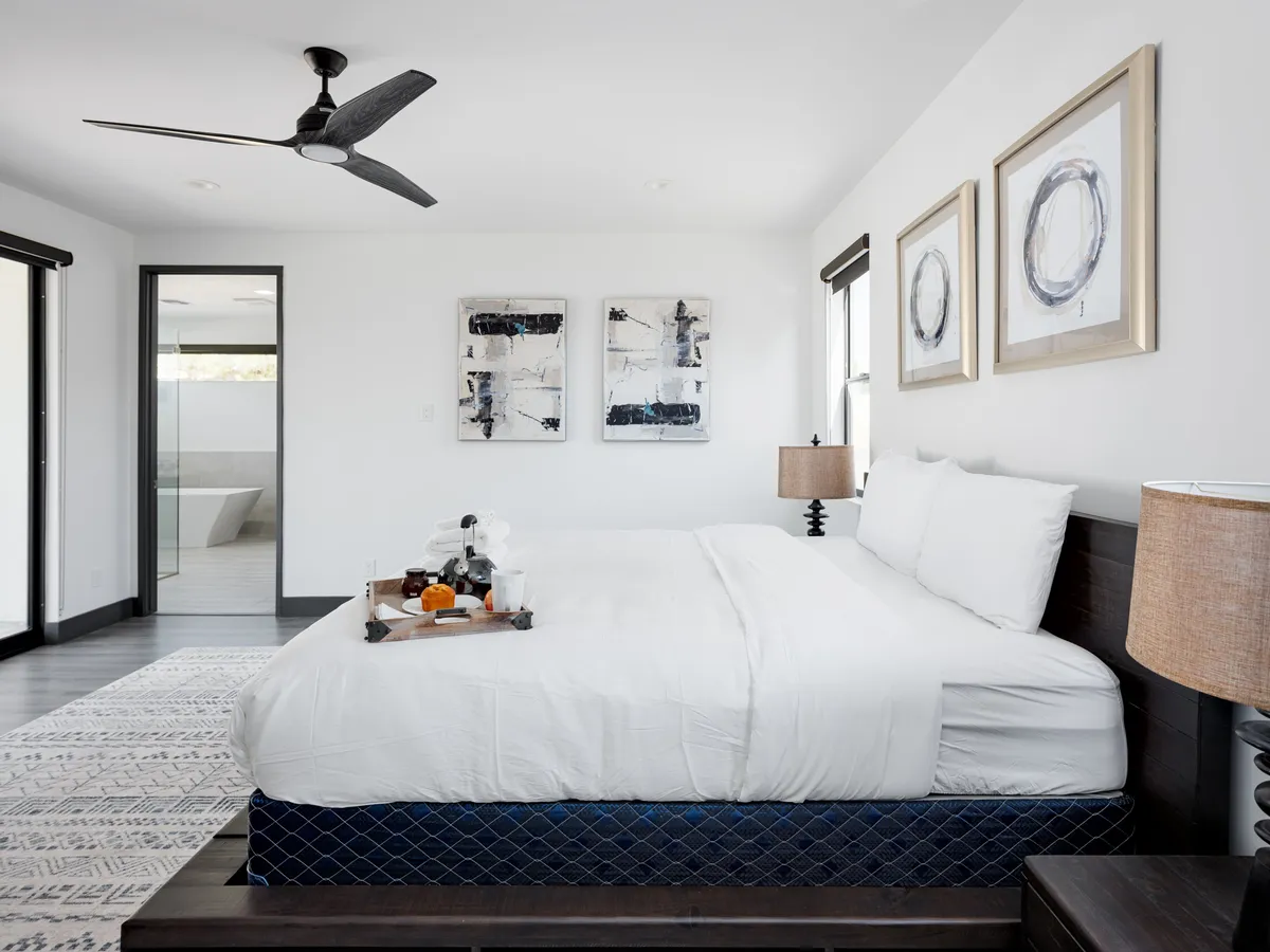A serene bedroom featuring a large bed with crisp white linens, decorative paintings on the wall, nightstands with lamps, and an open doorway leading to an en-suite bathroom. Natural light bathes the room, emphasizing its tranquil ambiance and sophisticated decor.