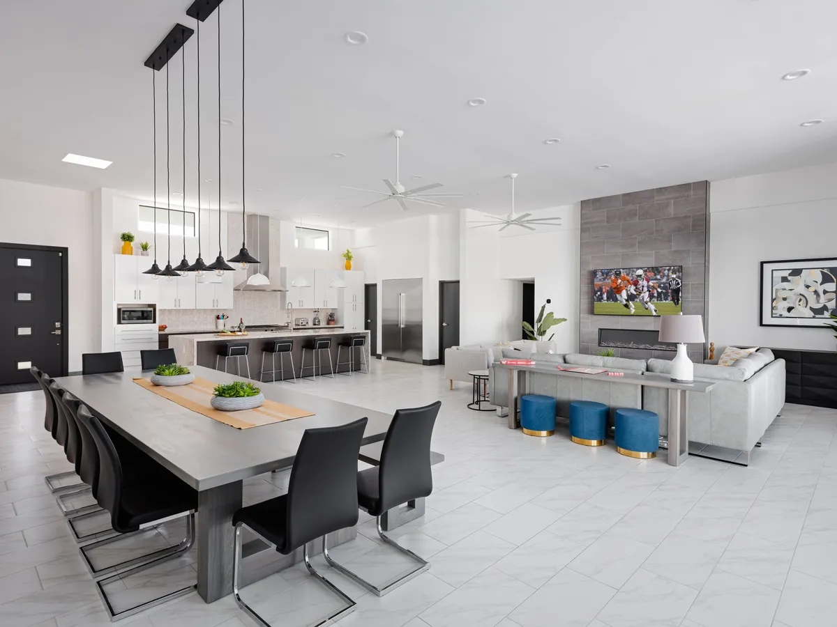 A wide view of a modern and inviting open-plan interior featuring a grand dining table set for a gathering, a cozy sofa area, a large kitchen island with seating, stylish lighting fixtures, and a television. The space is bathed in natural light and showcases designer furnishings with a blend of sophistication and comfort.