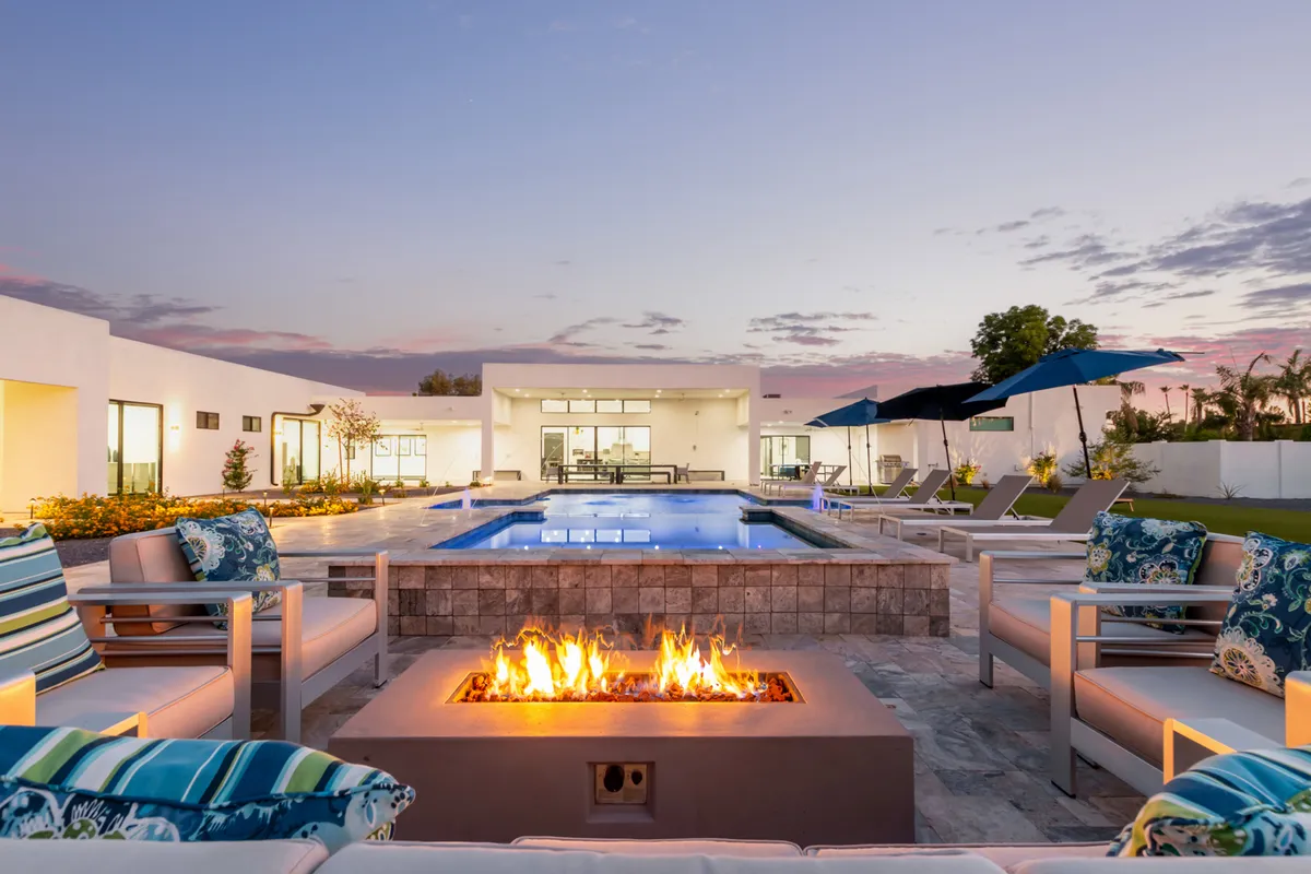 The hero image showcases the exterior of a modern Scottsdale mansion at sunset. The focal points include a sparkling swimming pool, a cozy fire pit surrounded by plush seating, and stylish outdoor umbrellas. The scene exudes a tranquil yet social atmosphere, inviting guests to unwind in luxury, enjoy intimate gatherings by the fire, and take in stunning desert sunset views.